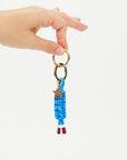 A hand holds The Upside LIMITLESS KEYRING, featuring gold metal hardware, a blue macrame paracord with a gold star charm, and two brown-tipped cord ends, shown against a plain white background.