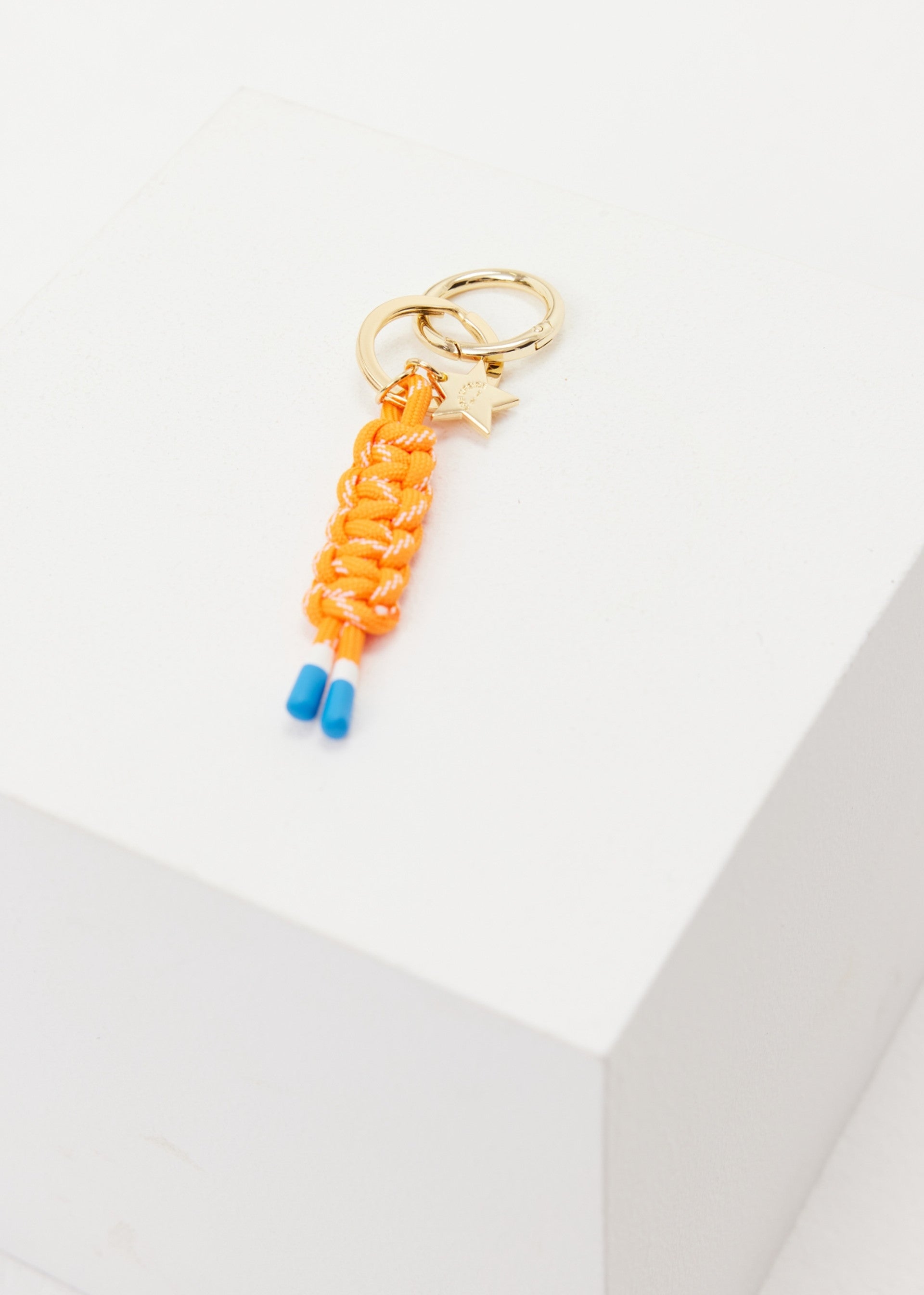 The Upside’s LIMITLESS KEYRING features a gold charm with a star and an orange macrame paracord braid, finished with blue tips, shown on a white geometric block.