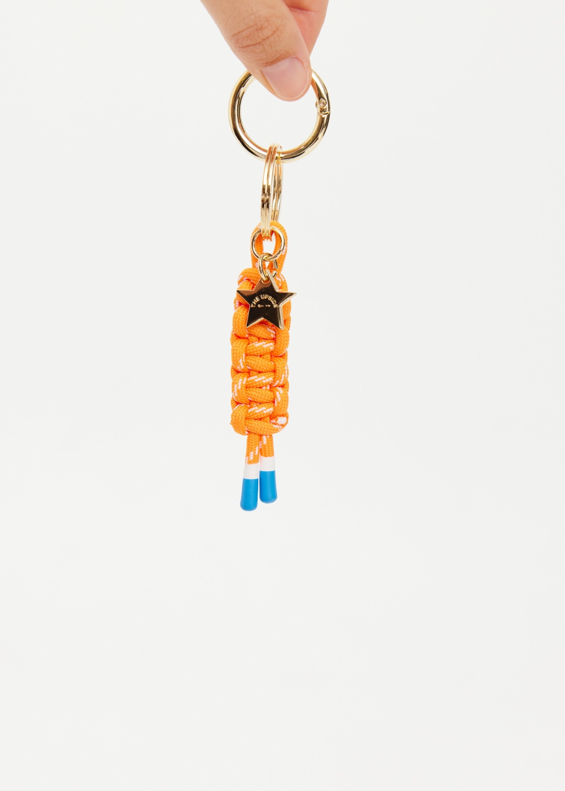 A hand holds The Upside's LIMITLESS KEYRING, a gold keyring with an orange macrame paracord keychain, blue-tipped ends, and a small metal star, shown against a plain white background.