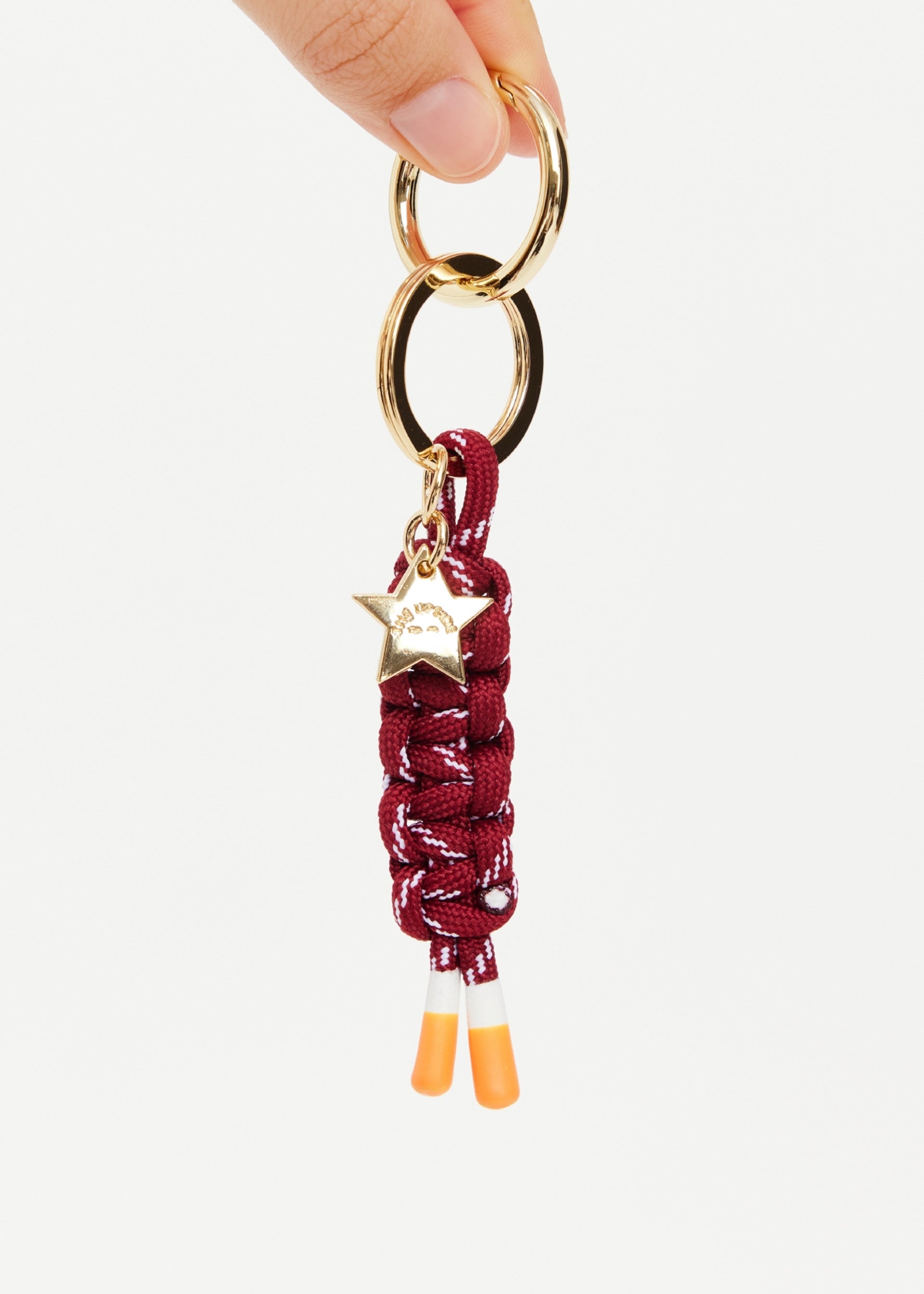 A hand displays The Upside LIMITLESS KEYRING, featuring maroon and white macrame paracord with orange-tipped ends and a gold star-shaped charm engraved with text.