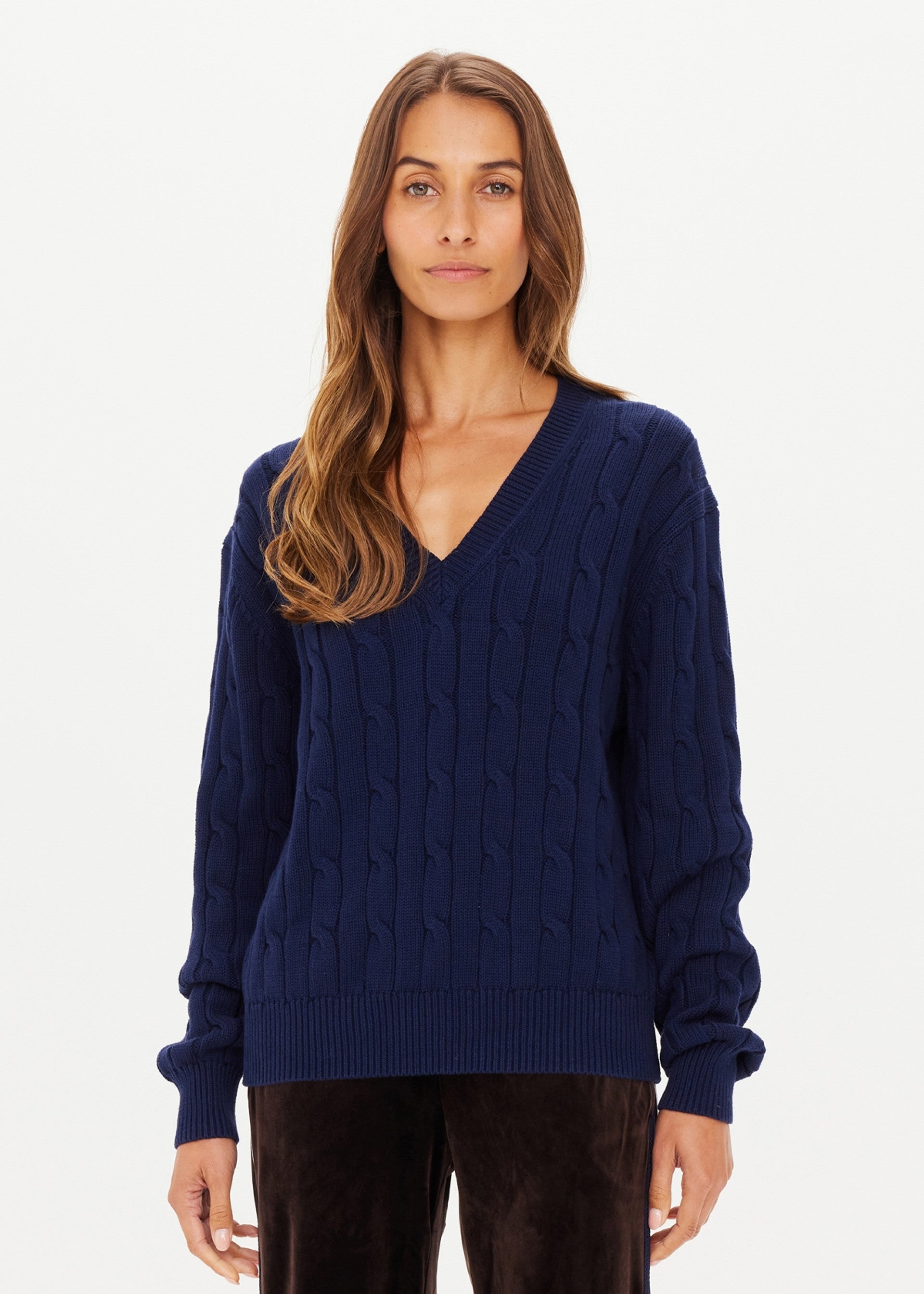 A woman with long brown hair wears The Upside’s SEYMOUR KNIT SWEATER, a navy blue cable-knit V-neck, paired with dark pants, standing against a plain white background.