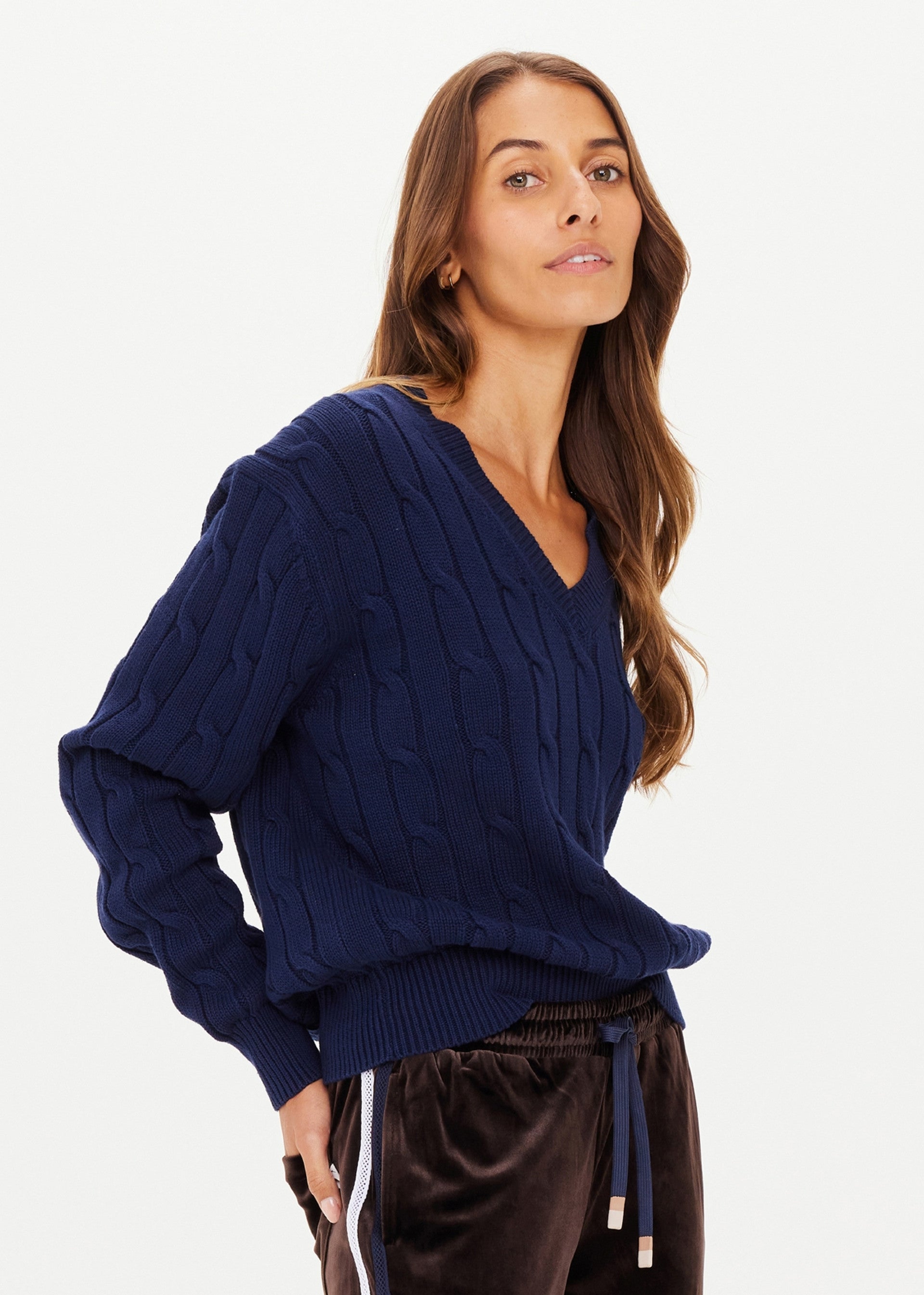 A woman with long brown hair stands confidently against a plain white background, wearing The Upside's SEYMOUR KNIT SWEATER—a navy blue cable-knit V-neck style—paired with dark brown velvet pants.