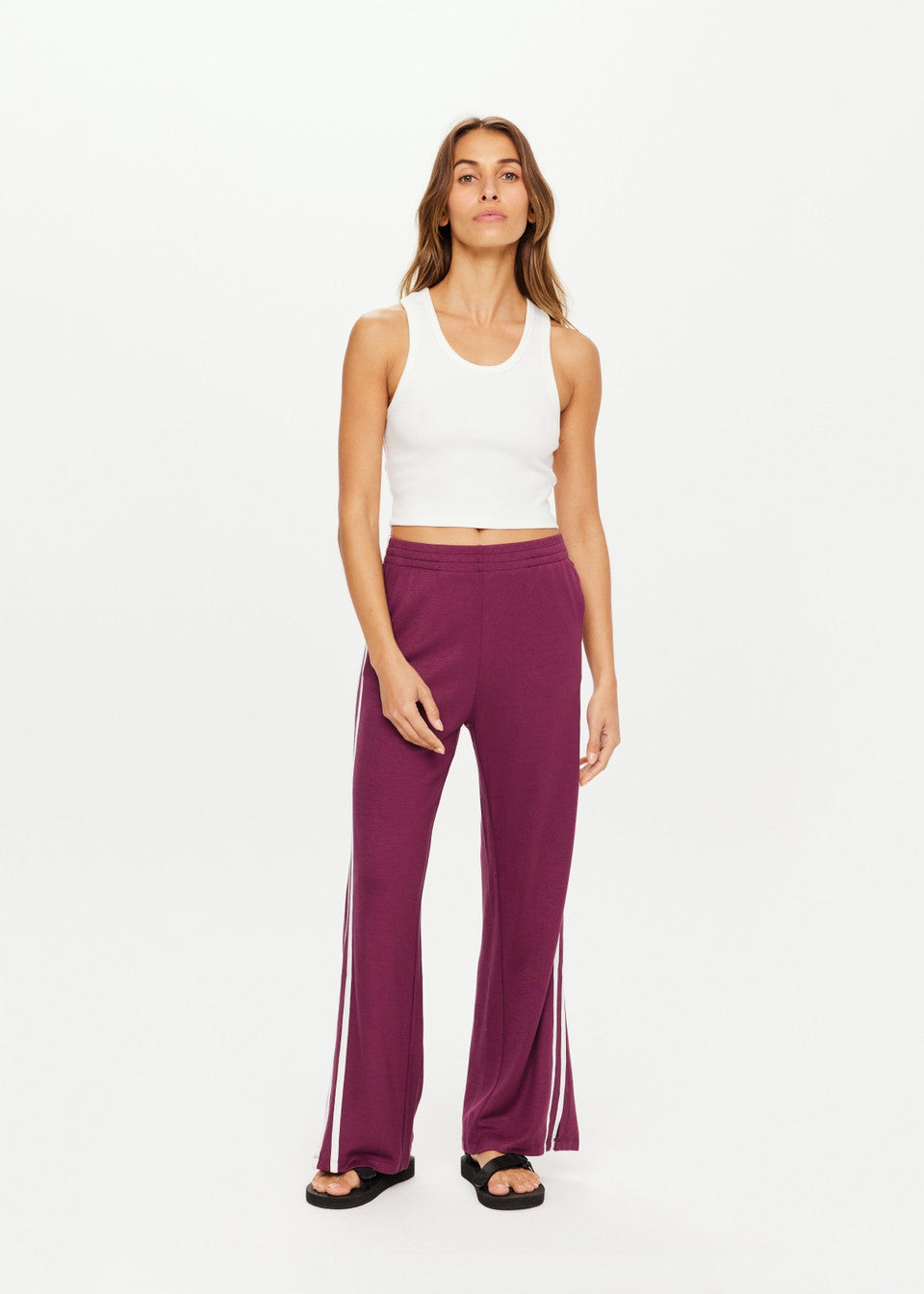 The woman stands confidently against a plain background, wearing The Upside's JULIET PANT—a stylish pair of purple low-rise pants with side stripes and an elastic waistband—and a white tank top. Her long, wavy hair cascades as she gazes directly at the camera with a neutral expression and sleek black sandals.