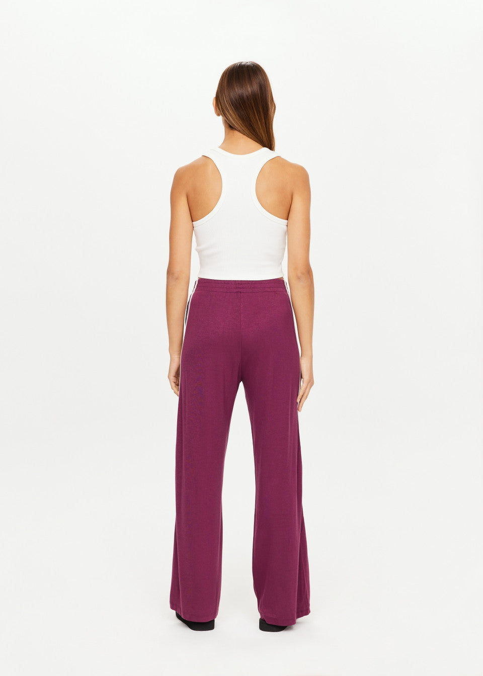 A woman with long brown hair, facing away from the camera, models The Upside's JULIET PANT. The purple wide-leg pants have a slinky fabric for elegance and an elasticated waist for comfort, paired with a white tank top against a plain white background.