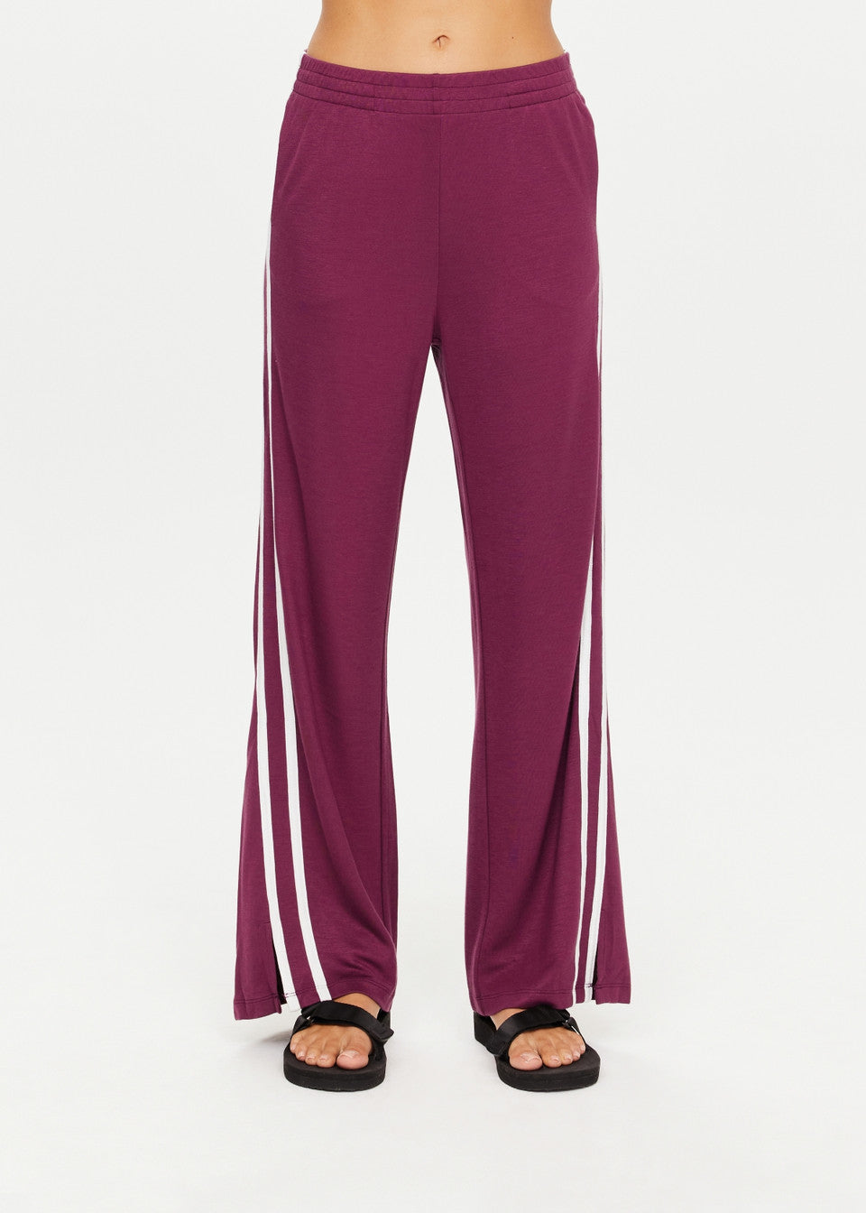 A person is wearing The Upside's JULIET PANT, purple wide-leg pants with white side stripes and an elastic waist, paired with black sandals. The slinky material of the pants enhances the smooth look against a plain white background.