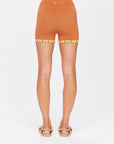 Wearing The Upside's VADELLA 4IN KNIT SPIN SHORT, a person is seen from the back in high-waisted, textured orange cotton shorts with a yellow and brown hem pattern. These playful shorts pair well with woven sandals against a plain white background.