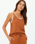 A woman showcases The Upside's ZAHARA JACQUIE TANK TOP, a brown sleeveless top with contrast stripe rib, paired with matching pants. Her long brown hair flows as she poses with hands in pockets against a plain white background, exuding retro basketball vibes.