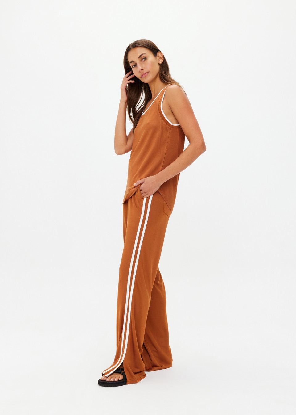 A person showcases a retro basketball vibe in The Upside's rust ZAHARA JACQUIE TANK TOP and matching wide-leg pants with contrast stripe ribs. They stand confidently on a neutral background, wearing black sandals and sporting long hair, looking towards the camera.