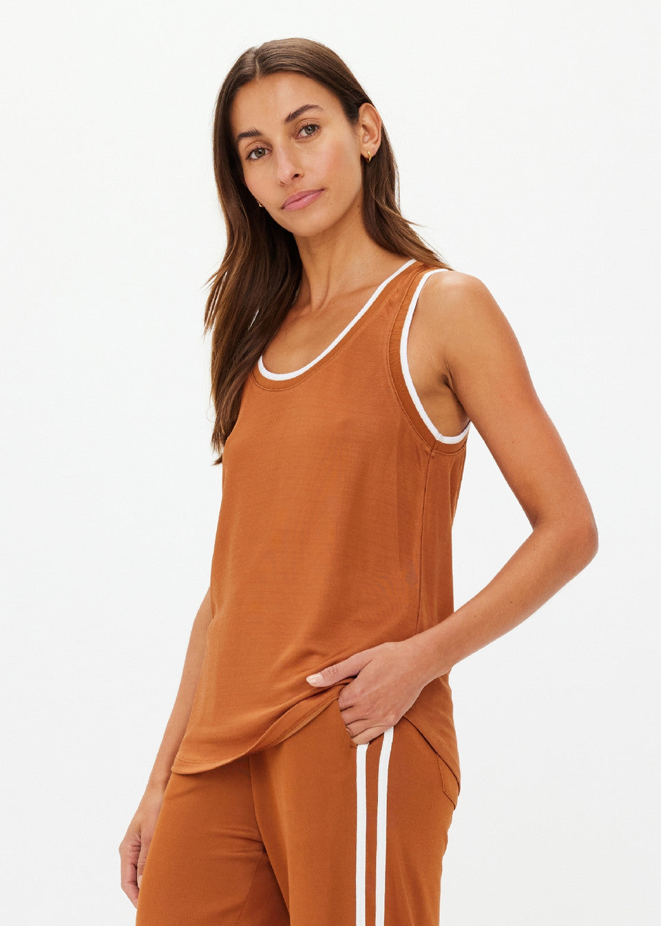 A person with long brown hair is wearing The Upside's ZAHARA JACQUIE TANK TOP and matching pants, showcasing a retro basketball vibe. They stand against a plain light background, gazing at the camera with a relaxed expression.