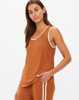 A person with long brown hair is wearing The Upside's ZAHARA JACQUIE TANK TOP and matching pants, showcasing a retro basketball vibe. They stand against a plain light background, gazing at the camera with a relaxed expression.