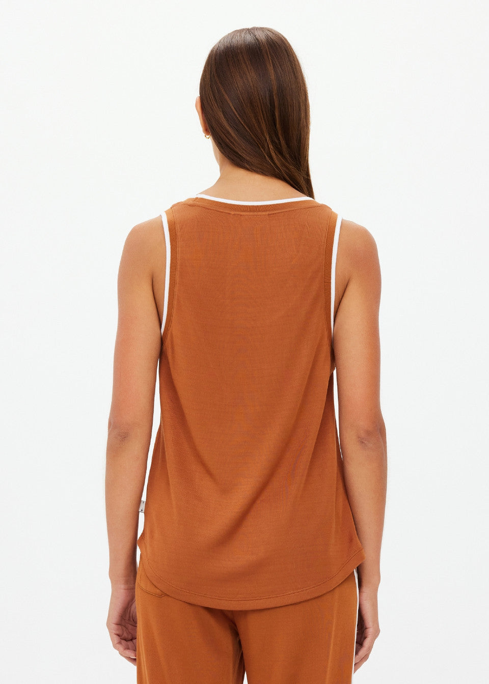 A person with long brown hair is wearing The Upside's ZAHARA JACQUIE TANK TOP, a sleeveless rust-colored top with contrast stripe rib. Facing away, they highlight the slinky back of the untucked top, paired with rust-colored pants against a plain white background.