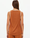 A person with long brown hair is wearing The Upside's ZAHARA JACQUIE TANK TOP, a sleeveless rust-colored top with contrast stripe rib. Facing away, they highlight the slinky back of the untucked top, paired with rust-colored pants against a plain white background.