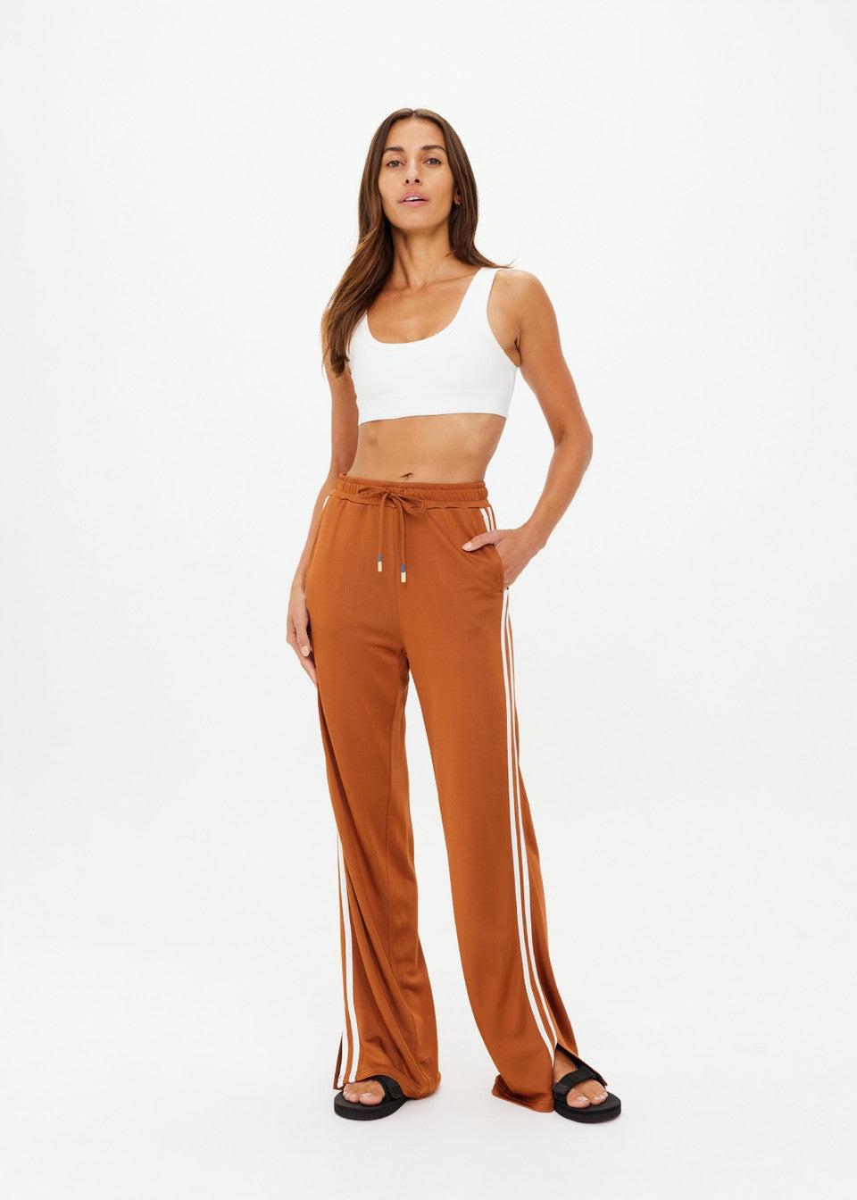 A person confidently wears The Upside's ZAHARA ROMA PANT, warm rust high-waisted with white side stripes, paired with a white crop top and black sandals. They have one hand in their pocket against a plain white background.