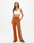 A person confidently wears The Upside's ZAHARA ROMA PANT, warm rust high-waisted with white side stripes, paired with a white crop top and black sandals. They have one hand in their pocket against a plain white background.