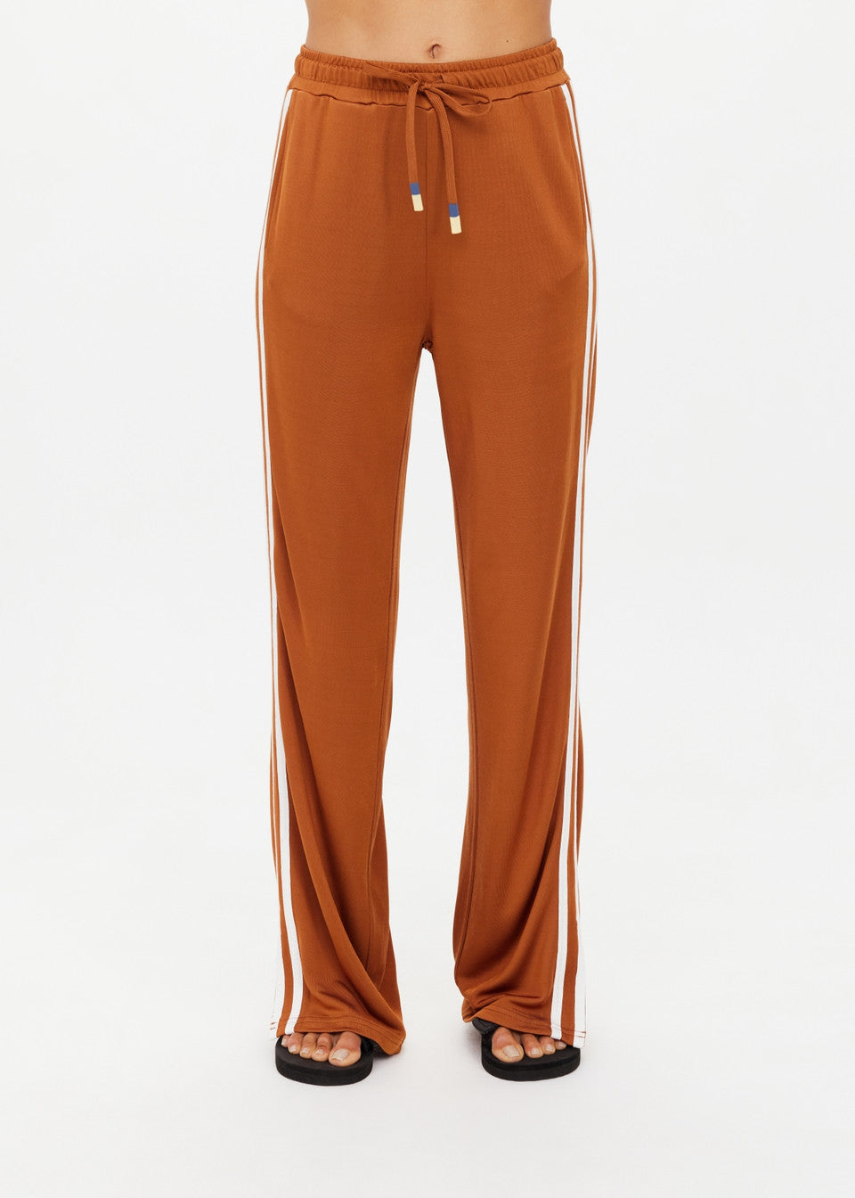 A person is wearing The Upside's ZAHARA ROMA PANT, high-waisted with an elasticated waist and white side stripes in a warm rust tone. Black flip-flops complete the look, with the photo cropped at the midsection to spotlight these stylish pants and casual footwear.