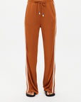 A person is wearing The Upside's ZAHARA ROMA PANT, high-waisted with an elasticated waist and white side stripes in a warm rust tone. Black flip-flops complete the look, with the photo cropped at the midsection to spotlight these stylish pants and casual footwear.