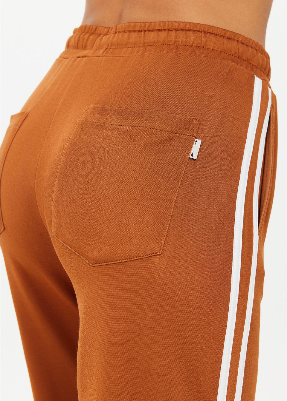 Close-up of a person in ZAHARA ROMA PANT by The Upside, featuring high-waisted brown sweatpants with a back pocket and white side stripes. The warm rust fabric looks soft and stretchy, highlighting the upper back of the pants.