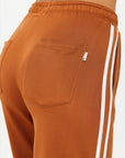 Close-up of a person in ZAHARA ROMA PANT by The Upside, featuring high-waisted brown sweatpants with a back pocket and white side stripes. The warm rust fabric looks soft and stretchy, highlighting the upper back of the pants.