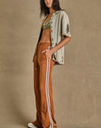 Against a plain backdrop, a person wears The Upside's beige patterned shirt, a warm rust bralette, ZAHARA ROMA PANT (brown high-waisted wide-leg pants with white side stripes), complemented by long dark hair and open-toed sandals.