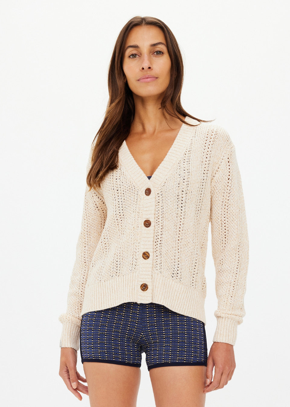 A woman with long brown hair wears The Upside's cream-colored DREW BUTTON KNIT CARDIGAN crafted from organic cotton and blue patterned shorts. Her relaxed fit attire complements her neutral expression as she stands against a plain white background.