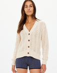 A woman with long brown hair wears The Upside's cream-colored DREW BUTTON KNIT CARDIGAN crafted from organic cotton and blue patterned shorts. Her relaxed fit attire complements her neutral expression as she stands against a plain white background.