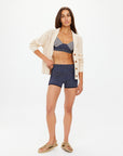 With long hair and a confident stance, the individual dons The Upside's DREW BUTTON KNIT CARDIGAN in cream over a patterned bikini top and high-waisted shorts, finishing the look with beige sandals against a plain white backdrop.