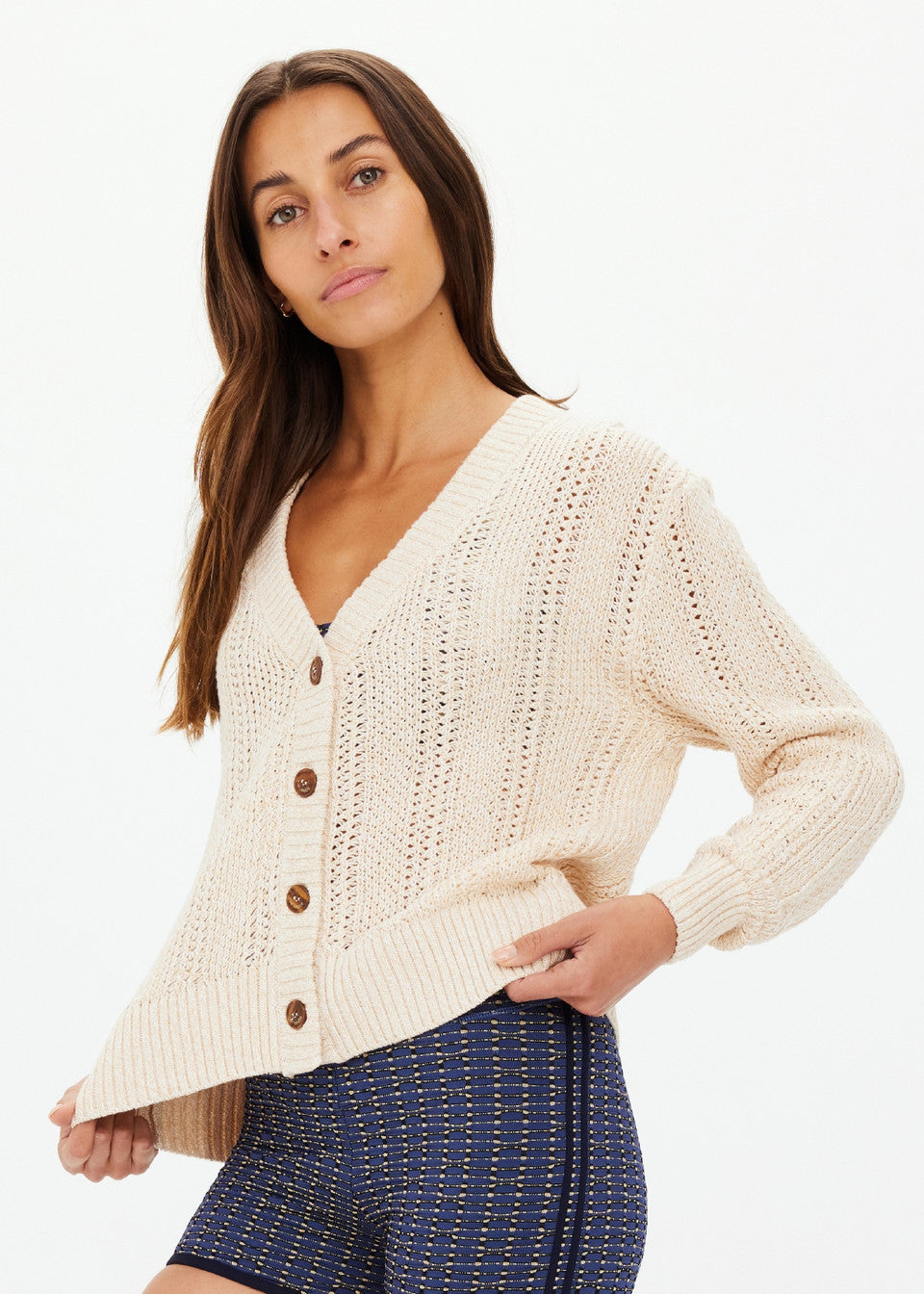 A person with long hair wearing The Upside's cream-colored DREW BUTTON KNIT CARDIGAN with tortoiseshell buttons and blue patterned shorts stands against a plain background, confidently looking at the camera.