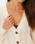 A woman wears The Upside's DREW BUTTON KNIT CARDIGAN, a cream-colored relaxed fit with tortoiseshell buttons, over a blue top. Her long brown hair drapes over one shoulder as she touches a button, with her head partially out of the frame.