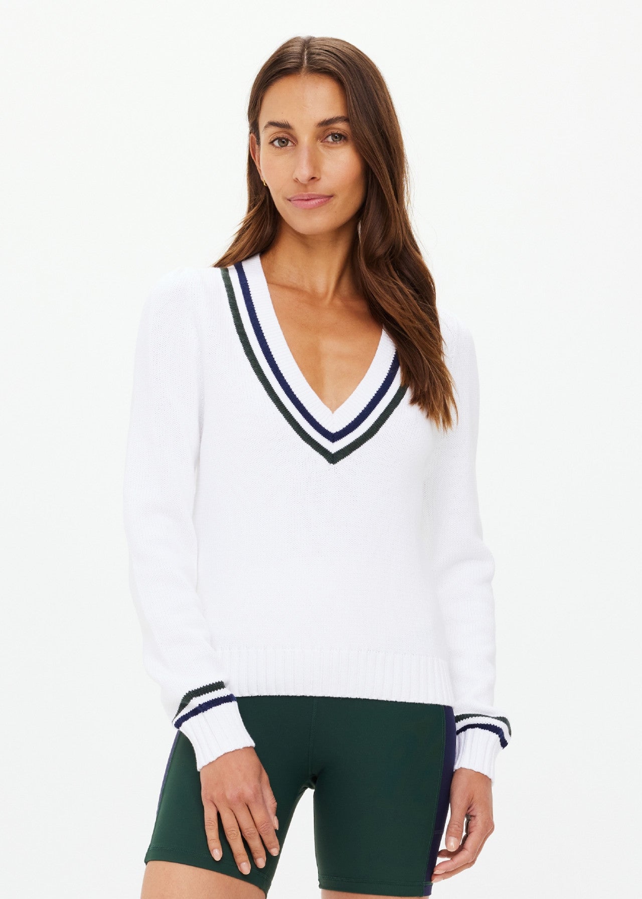 A person with long brown hair wears The Upside's SONNY V NECK KNIT SWEATER made from organic cotton, featuring dark striped edges and paired with dark green shorts. An embroidered arrow logo accents their look against a plain white background.