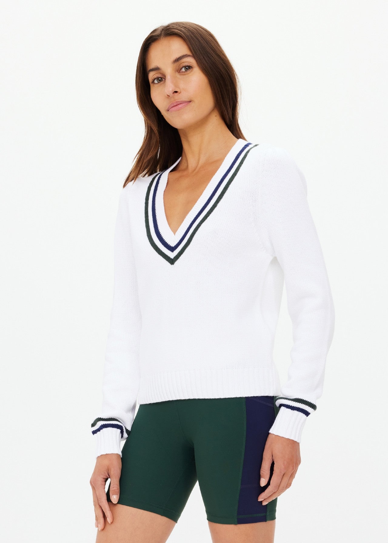 A person with long brown hair models The Upside's SONNY V NECK KNIT SWEATER, white with black stripes and an embroidered arrow logo, paired with green and navy organic cotton cycling shorts, against a plain white background.