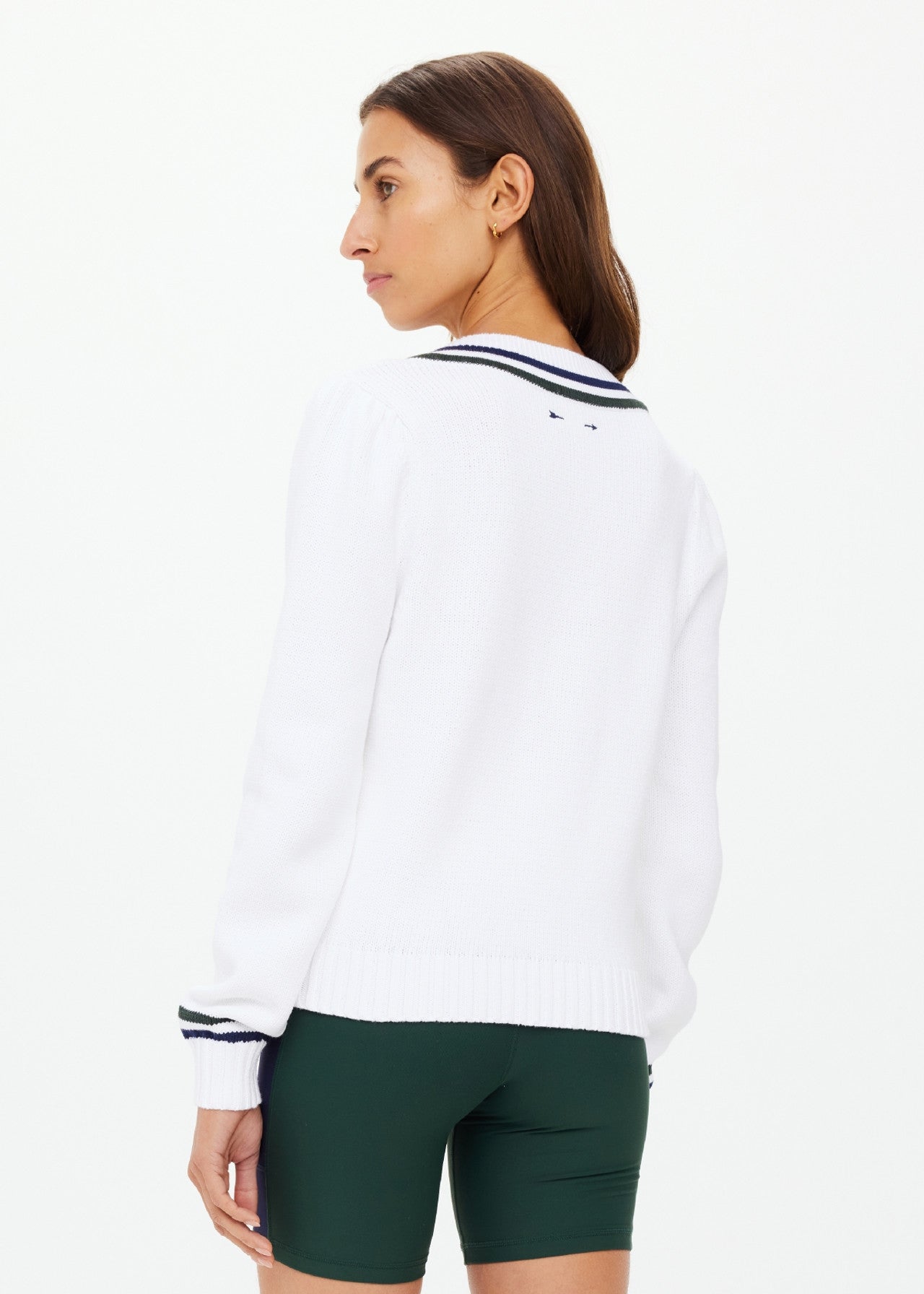 Someone stands and looks to the side, wearing The Upside's white SONNY V NECK KNIT SWEATER with black trim and green shorts. The organic cotton piece features an embroidered arrow logo, subtly enhancing its style against a plain white background.