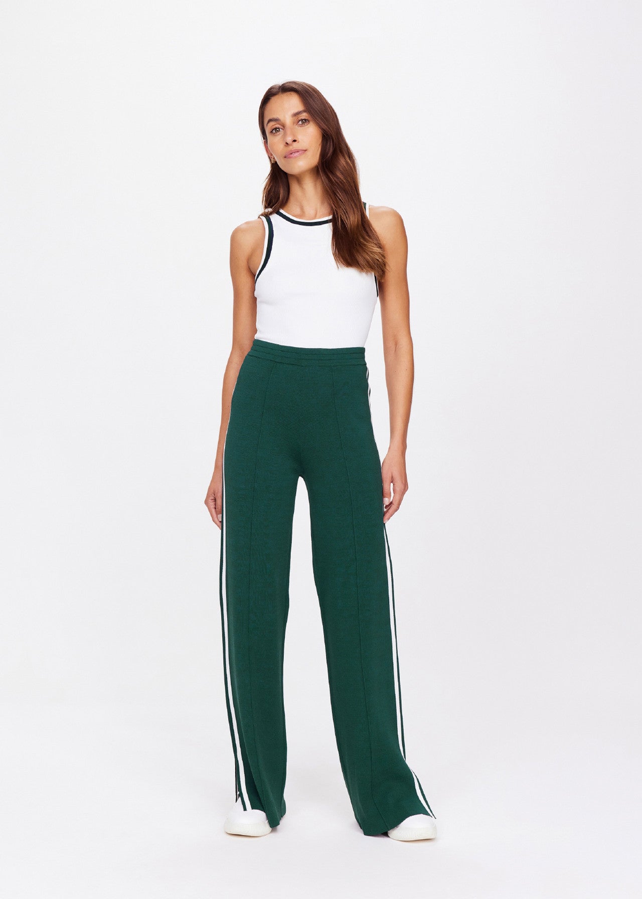 A person is wearing The Upside's LES BORDER FREYA KNIT PANT, a green straight-leg style with an elasticated waistband and white side stripes, paired with a white sleeveless top. They have long dark hair and pose against a plain light background.