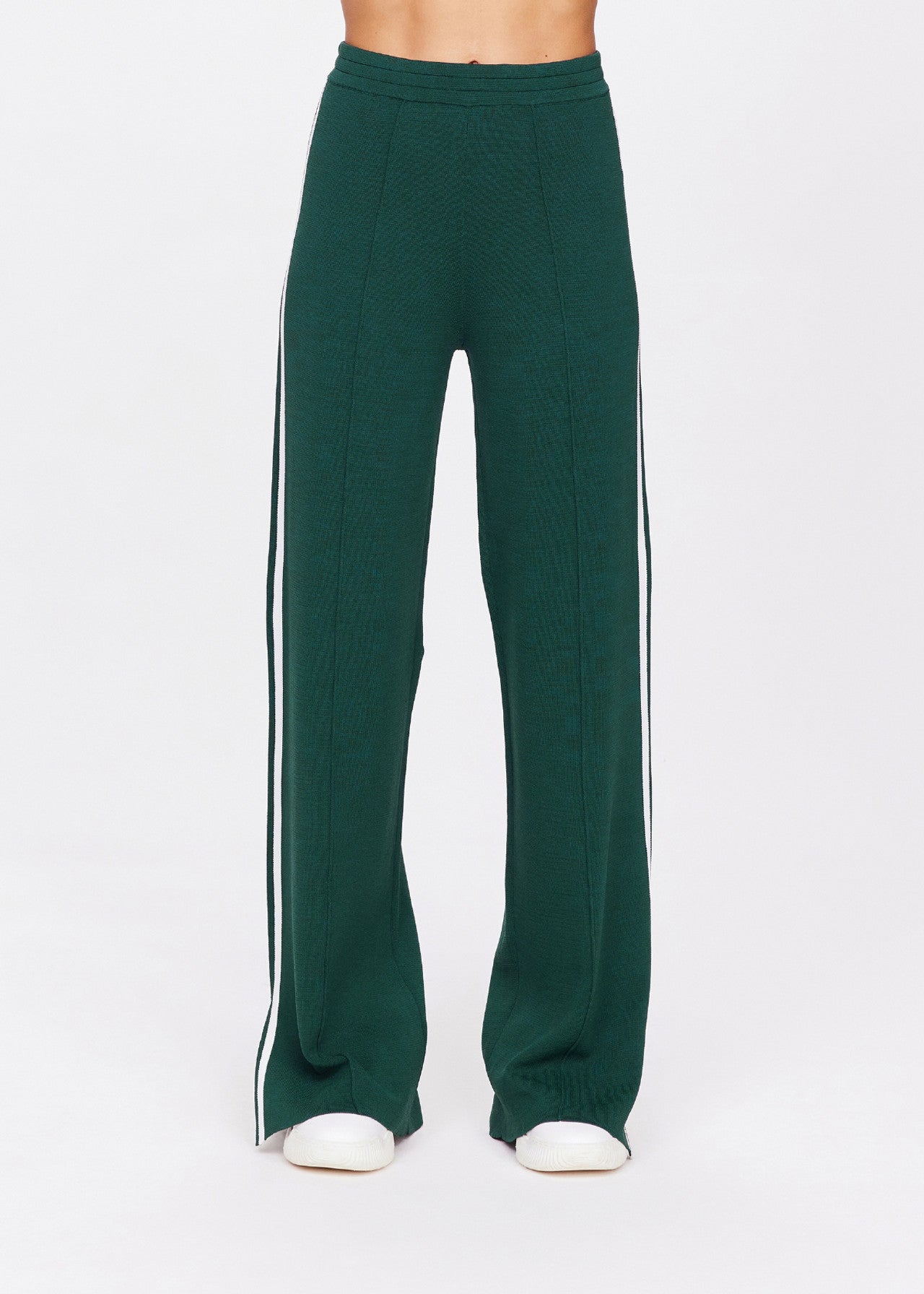 A person models The Upside's LES BORDER FREYA KNIT PANT, featuring green wide-leg design with white vertical stripes and an elastic waistband. Embroidered tonal arrow logo is visible above white shoes against a white background. Upper body not shown.