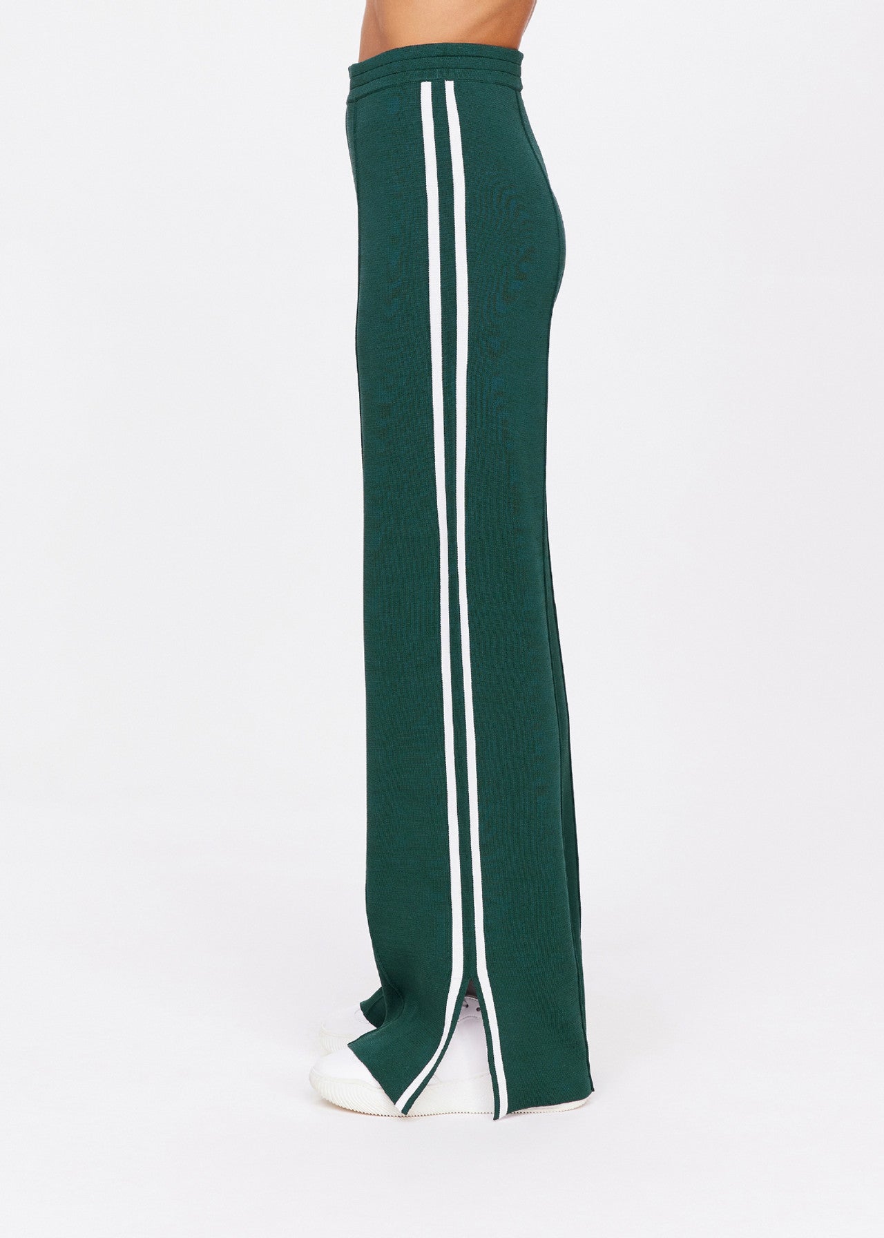 A person models The Upside's LES BORDER FREYA KNIT PANT, showcasing its green hue, white side stripes, and bottom slit. The pants feature an elasticated waistband and are presented in a side view on a plain white background.