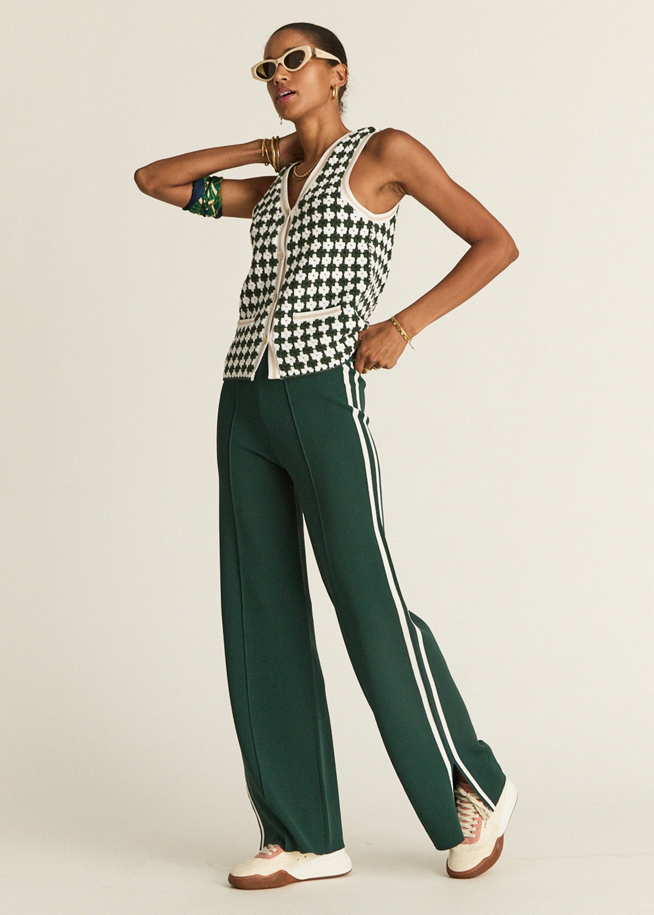 A person in sunglasses poses confidently, wearing The Upside's LES BORDER FREYA KNIT PANT and a black-and-white houndstooth vest, paired with white sneakers. They accessorize with a green bracelet and gold jewelry against a plain background.