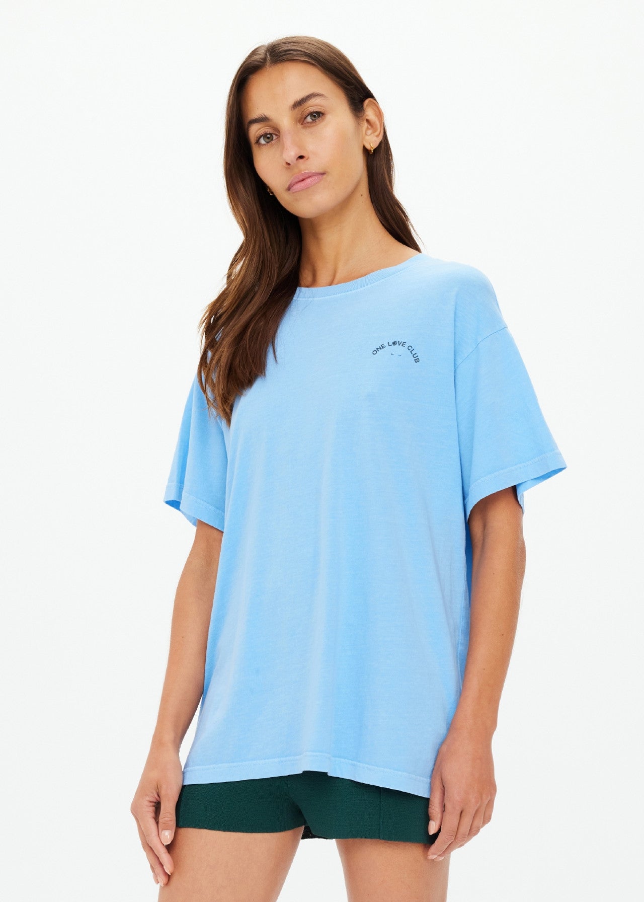 A person with long hair wears The Upside's ONE LOVE CLUB SAMMI TEE in sky blue and dark green shorts, standing against a white background. They have a relaxed posture with one hand by their side, and small text is visible on the organic cotton tee near the shoulder.