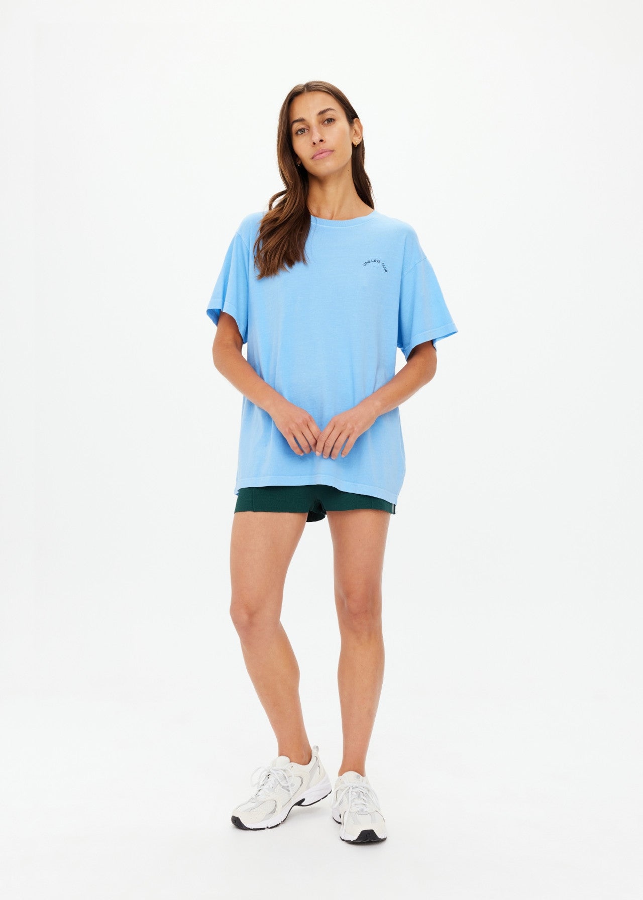 A woman poses gracefully against a plain backdrop, wearing The Upside's ONE LOVE CLUB SAMMI TEE in sky blue, paired with green shorts and white sneakers. Her long brown hair cascades beautifully as she clasps her hands in front.