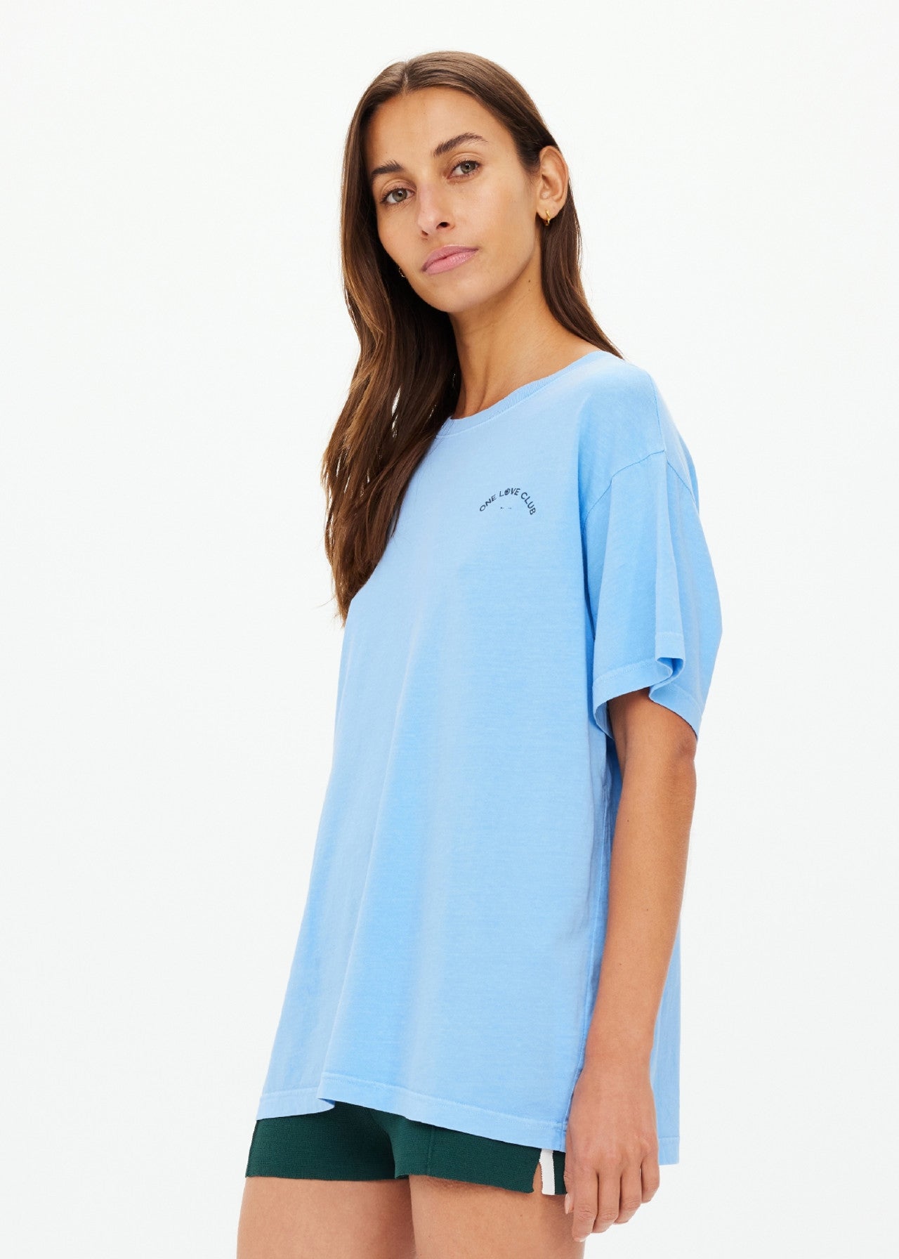 A person with long hair wears The Upside's sky-blue ONE LOVE CLUB SAMMI TEE, made from organic cotton, paired with dark green shorts. They're standing against a plain white background, confidently looking at the camera.