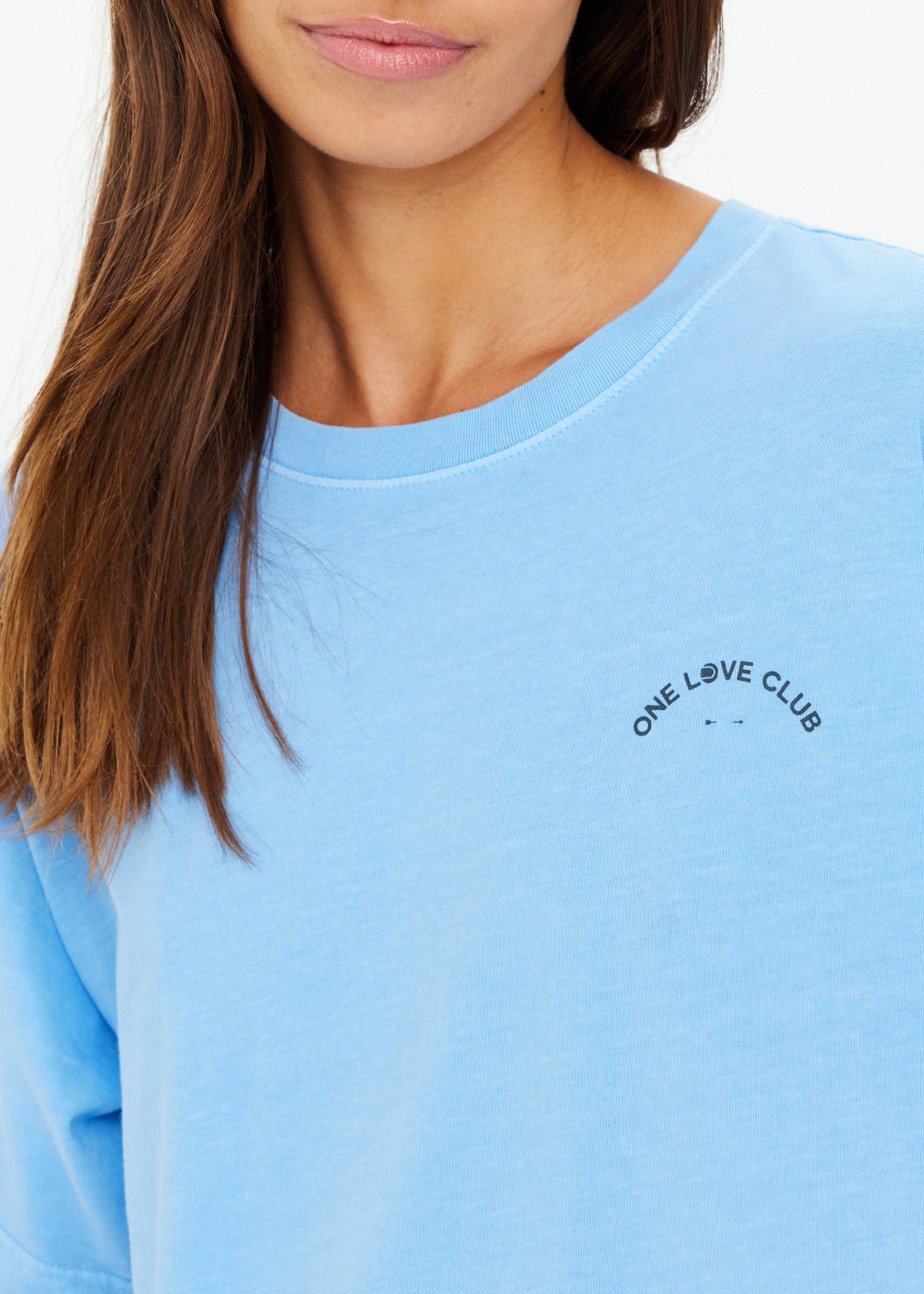 A woman wears The Upside's ONE LOVE CLUB SAMMI TEE in an oversized sky blue style, featuring the text "ONE LOVE CLUB" on the chest. Her straight brown hair falls over her shoulder, highlighting the organic cotton against the simple white backdrop.