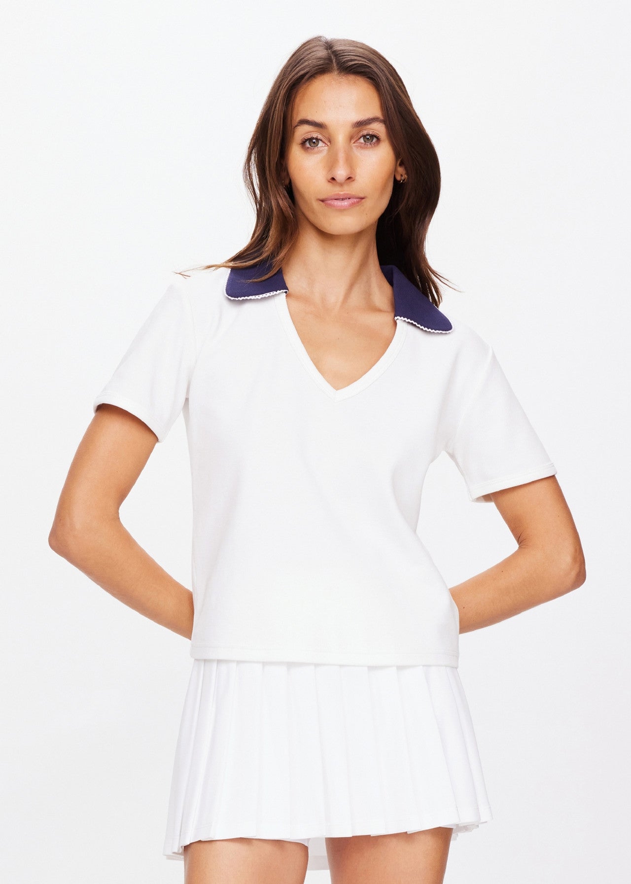 A person with long hair wears The Upside's JACOBS V NECK COLLARED POLO—a slim-fit white short-sleeved top with a dark collar and an embroidered logo—paired with a white pleated skirt. They stand against a plain white background, arms relaxed by their sides.