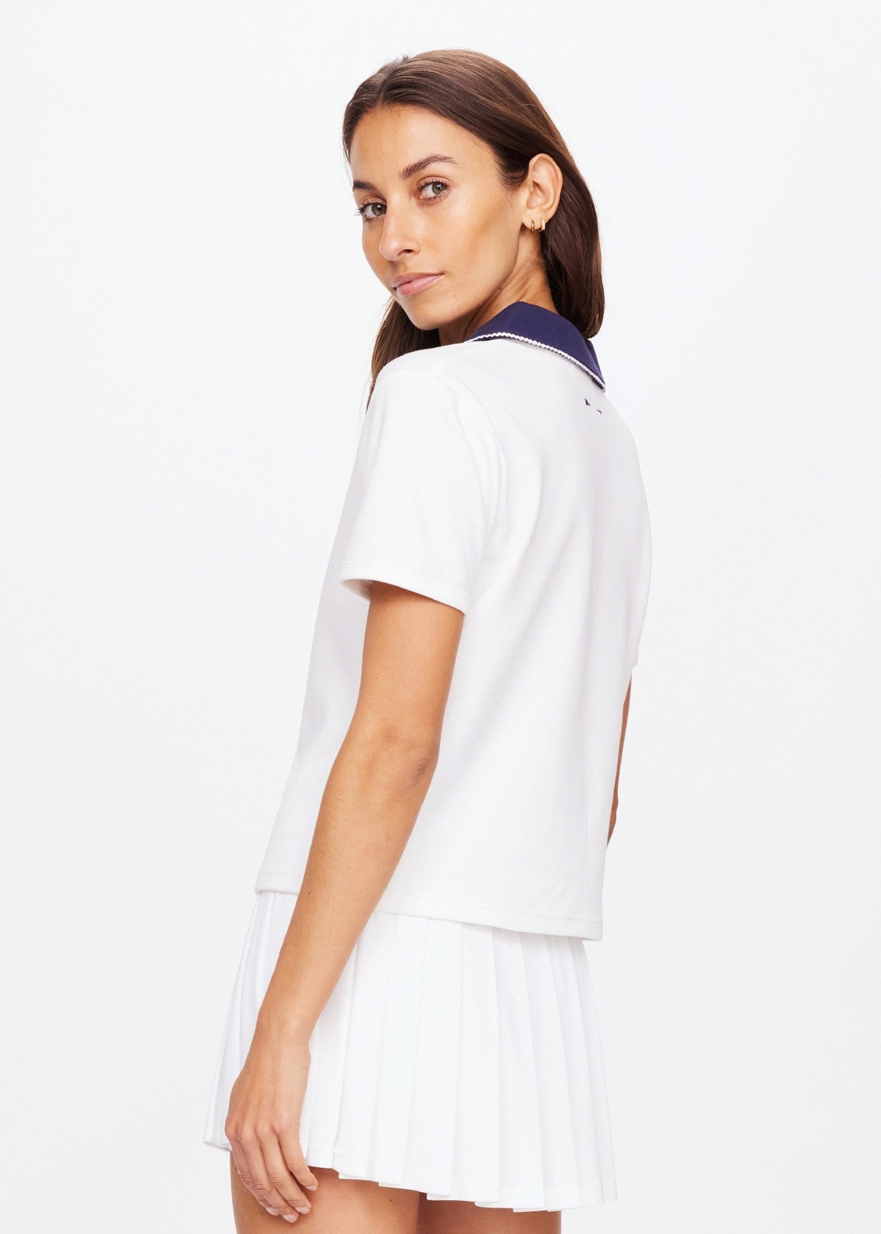 A person with long hair wears The Upside's JACOBS V NECK COLLARED POLO and a white pleated skirt, looking over their shoulder against a plain white background.