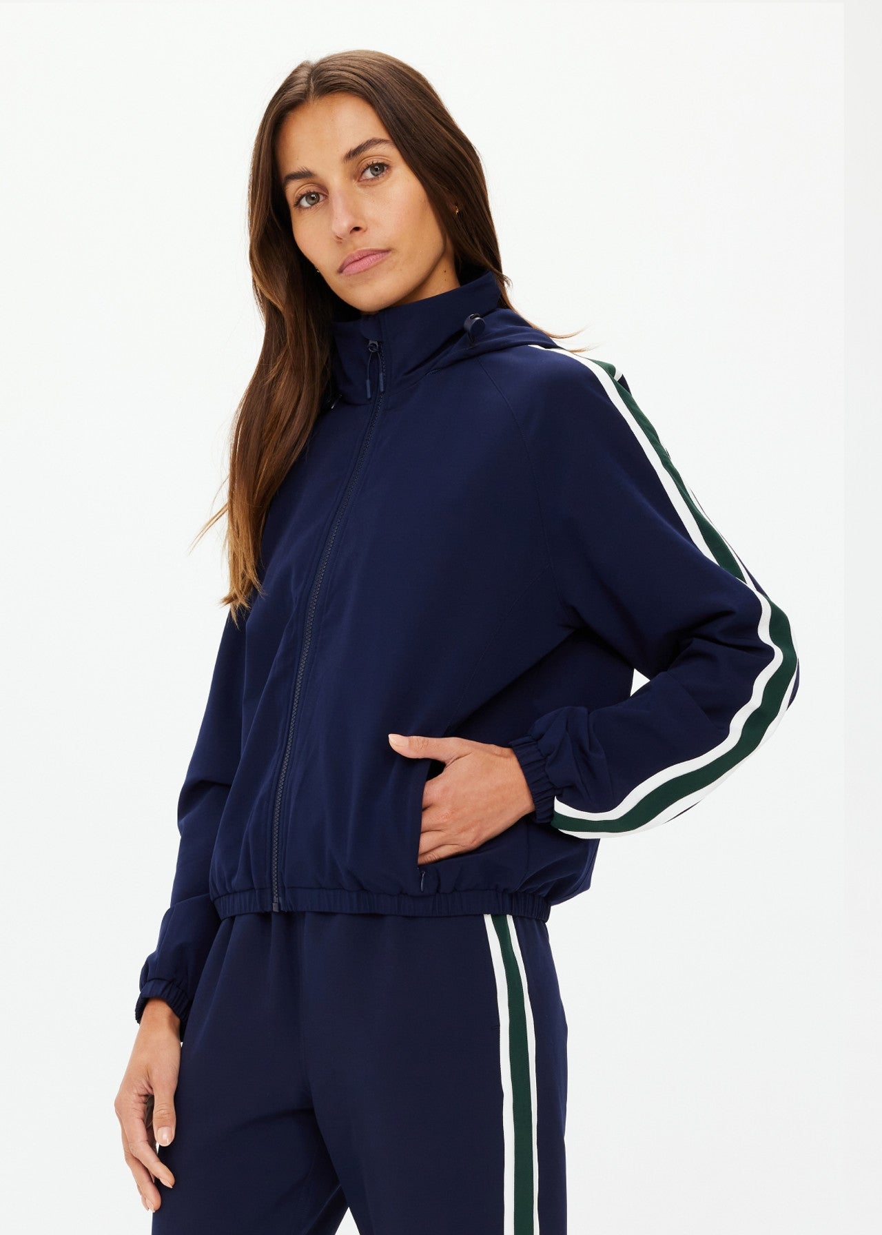 A person stands against a white background wearing The Upside's MANSI DAWSON RUN JACKET NAVY, featuring recycled polyester, a hood, and zippered front. The jacket has white and green stripes on the sleeves and pants. They look at the camera with a neutral expression.