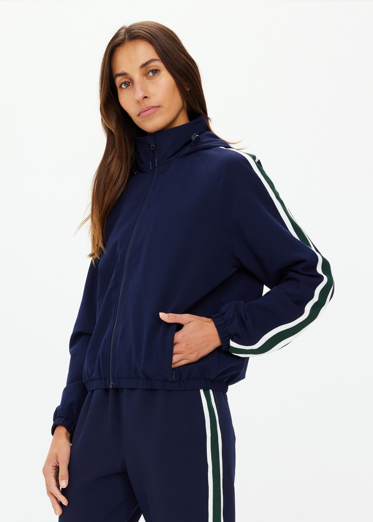 A person with long brown hair and a neutral expression wears The Upside's MANSI DAWSON RUN JACKET NAVY, featuring white and green striped sleeves, water-resistant recycled polyester, standing against a plain background with one hand on their hip.