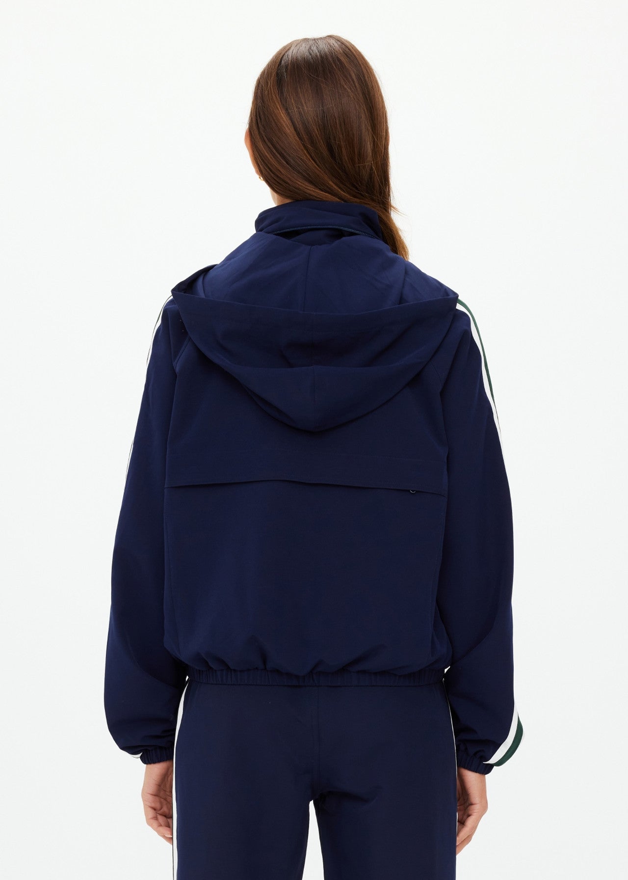 A person with long brown hair is seen from behind, wearing The Upside's MANSI DAWSON RUN JACKET NAVY, made from recycled polyester with white-striped sleeves and matching pants. This water-resistant ensemble stands out against the plain white background.