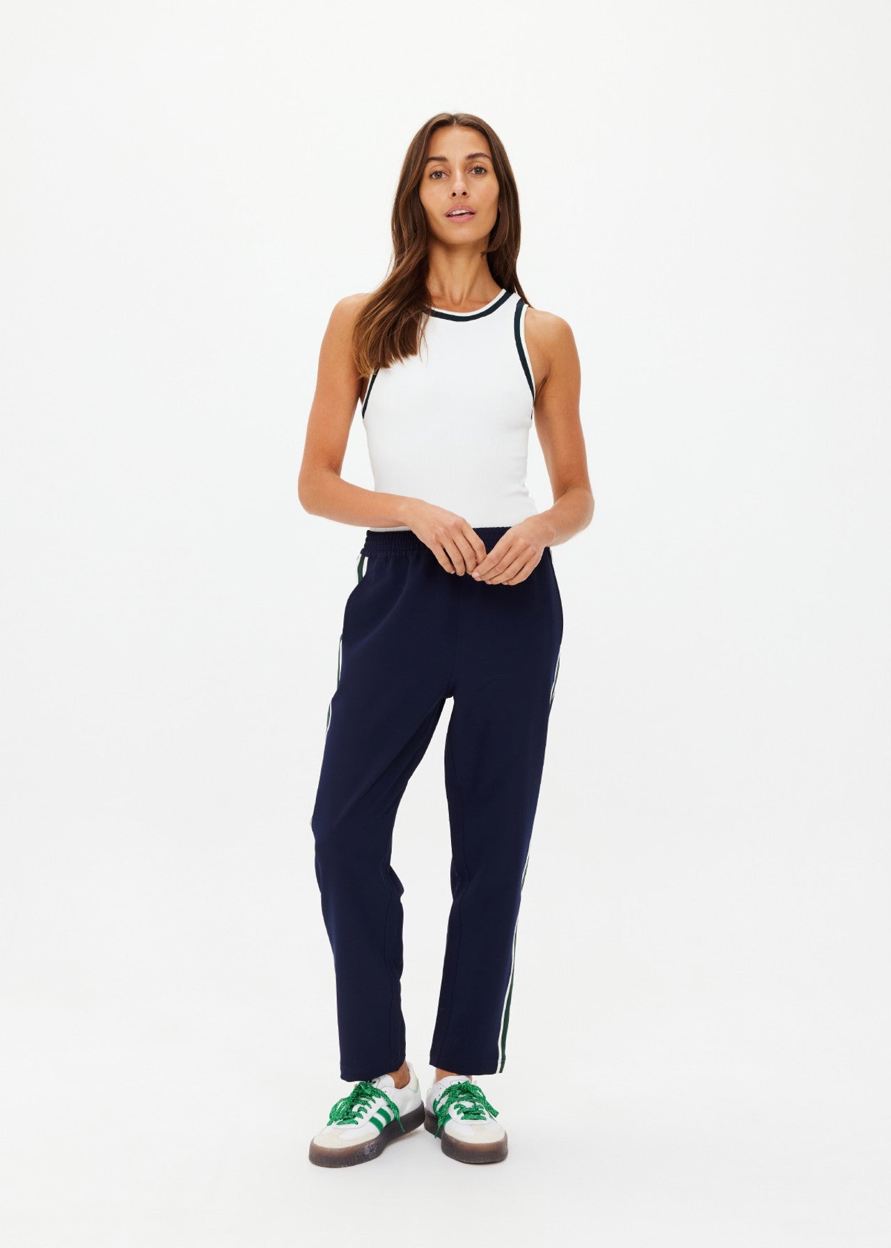 A person with long brown hair is wearing a white sleeveless top made from recycled polyester and The Upside's Mansi Paxton Tapered Pant in dark blue with side stripes. They stand against a plain white background, sporting moisture-wicking green and white sneakers.