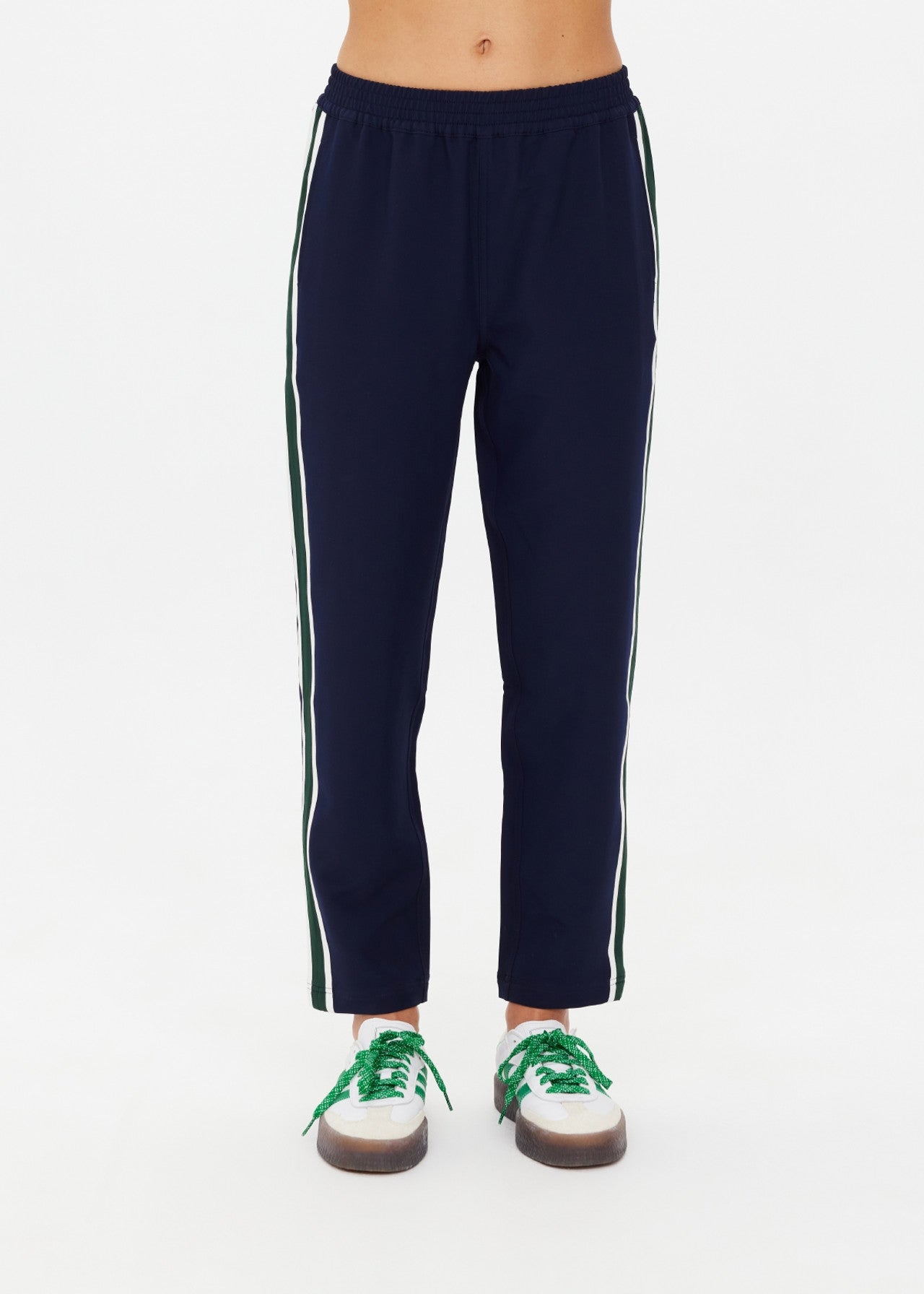 Someone wearing The Upside's MANSI PAXTON TAPERED PANT in navy with white and green side stripes, made from moisture-wicking recycled polyester, paired with white sneakers featuring green laces, stands against a plain white background.