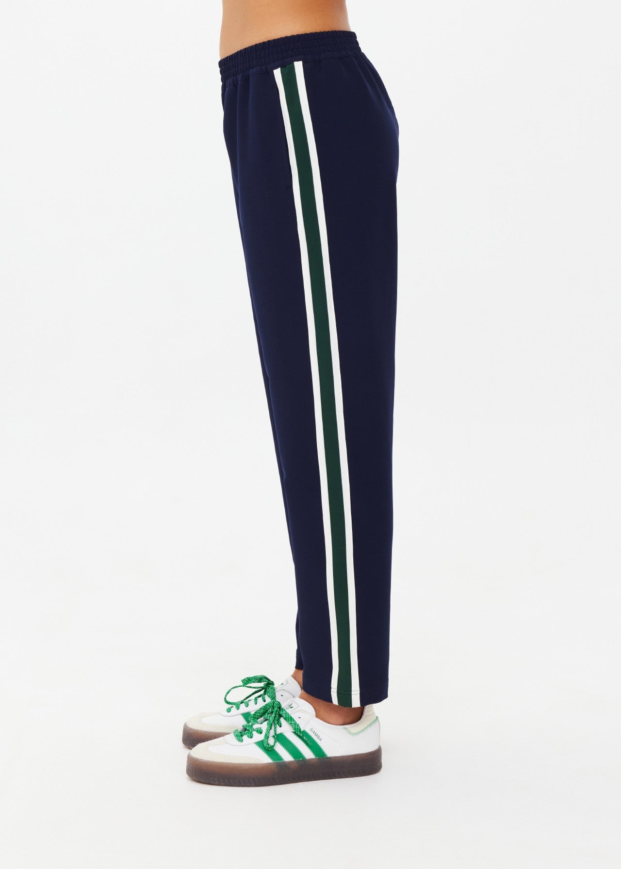 A person models The Upside's MANSI PAXTON TAPERED PANT, crafted from recycled polyester in navy blue with white and green stripes, teamed with white sneakers showcasing green accents. They stand in side profile against a plain white backdrop.