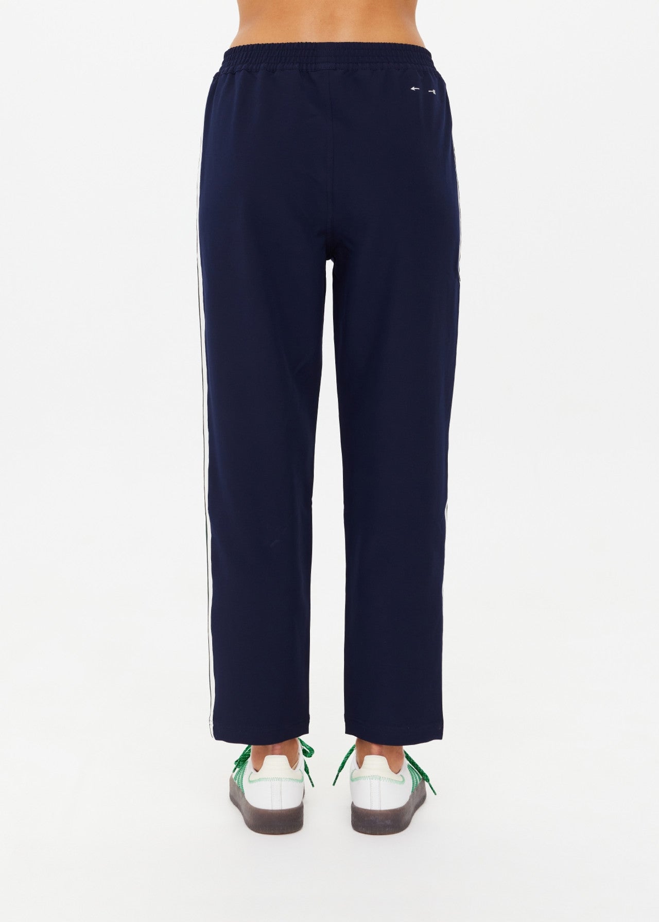 In the image, a person stands on a plain, light background, wearing The Upside's MANSI PAXTON TAPERED PANT in navy-blue with white side stripes crafted from recycled polyester. White sneakers with green accents complete their lower half attire.