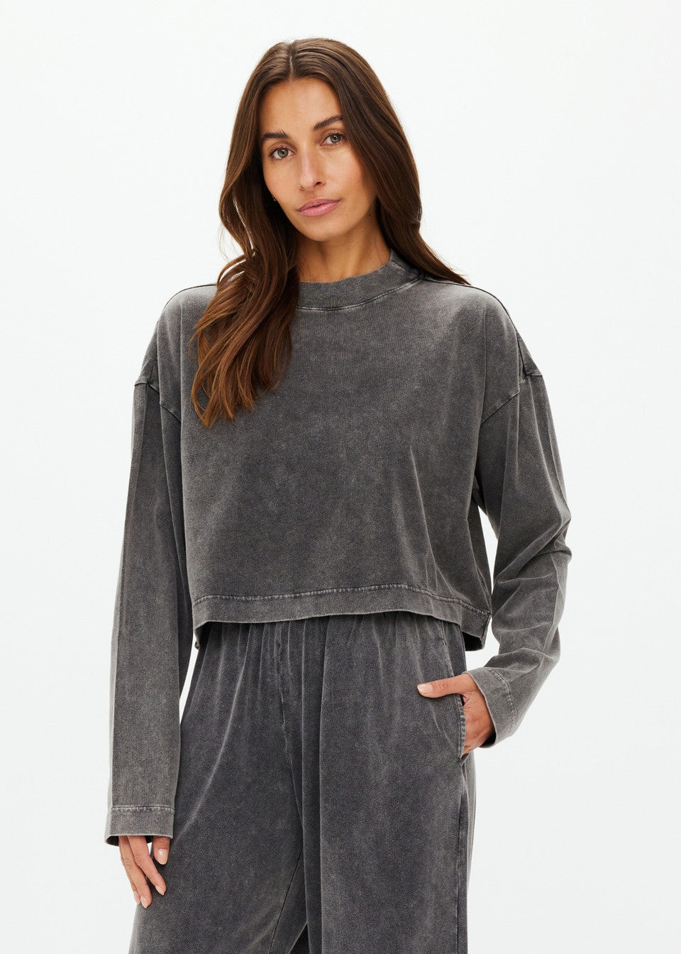 A woman poses in a relaxed demeanor wearing The Upside's charcoal grey Soho Cropped Long Sleeve Top and matching loose-fitting pants made from recycled cotton. She stands against a plain white background with one hand in her pocket.