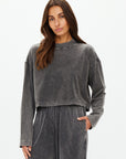 A woman poses in a relaxed demeanor wearing The Upside's charcoal grey Soho Cropped Long Sleeve Top and matching loose-fitting pants made from recycled cotton. She stands against a plain white background with one hand in her pocket.